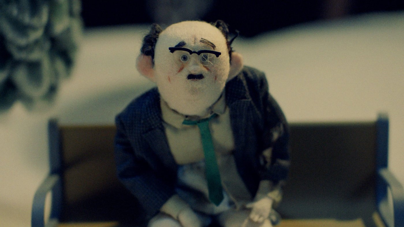 A felt stop-motion puppet of a balding man with glasses and a green tie sitting on a bench