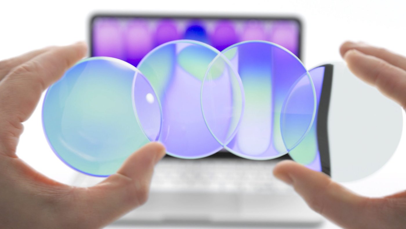 Two hands holding translucent glass discs in front of a laptop screen glowing purple and green