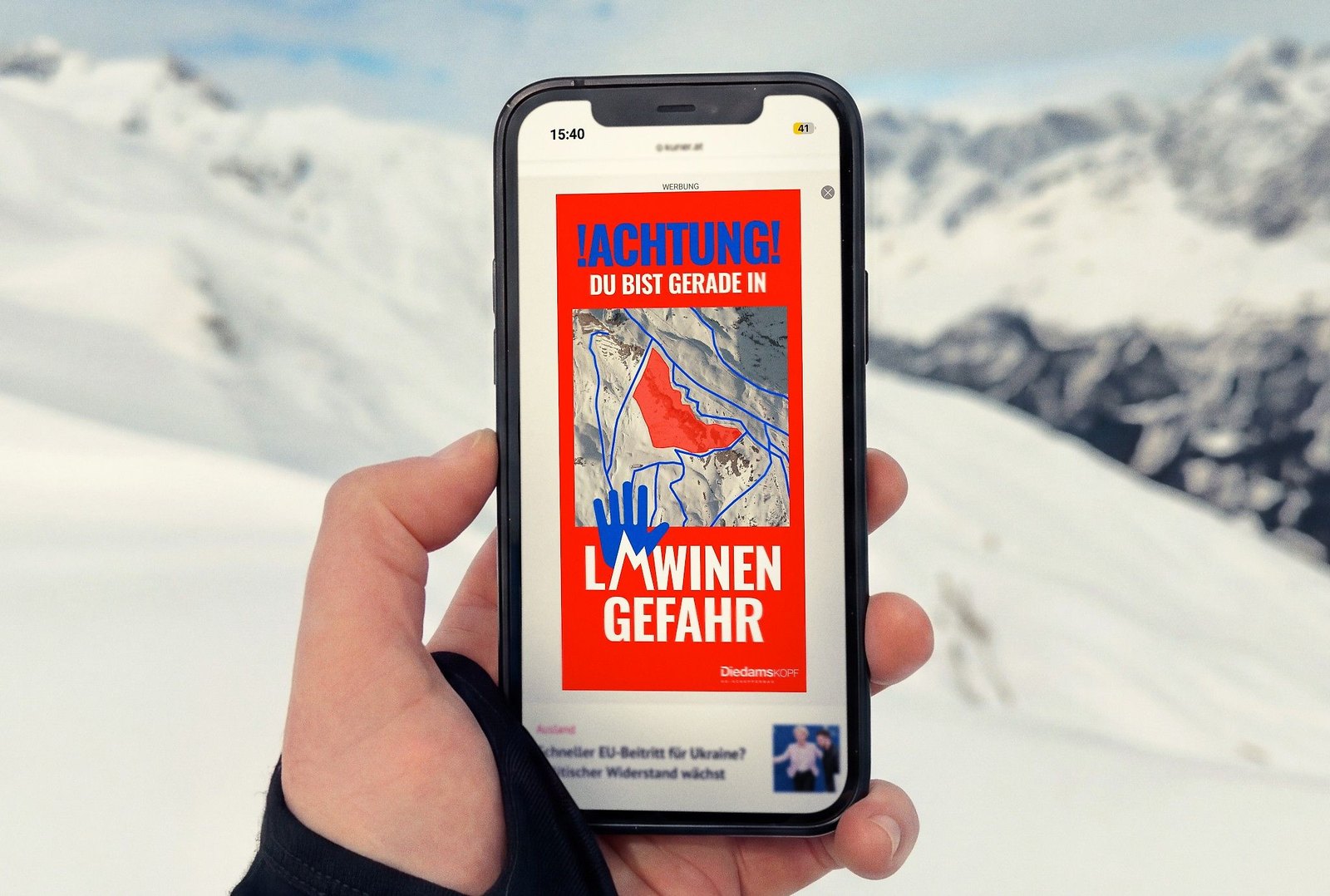 A hand holds a phone on a snowy mountain; on a news site a red vertical ad reads ACHTUNG, Du bist gerade in Lawinengefahr, around a map with the danger zone shaded red, with the Diedamskopf logo at the bottom