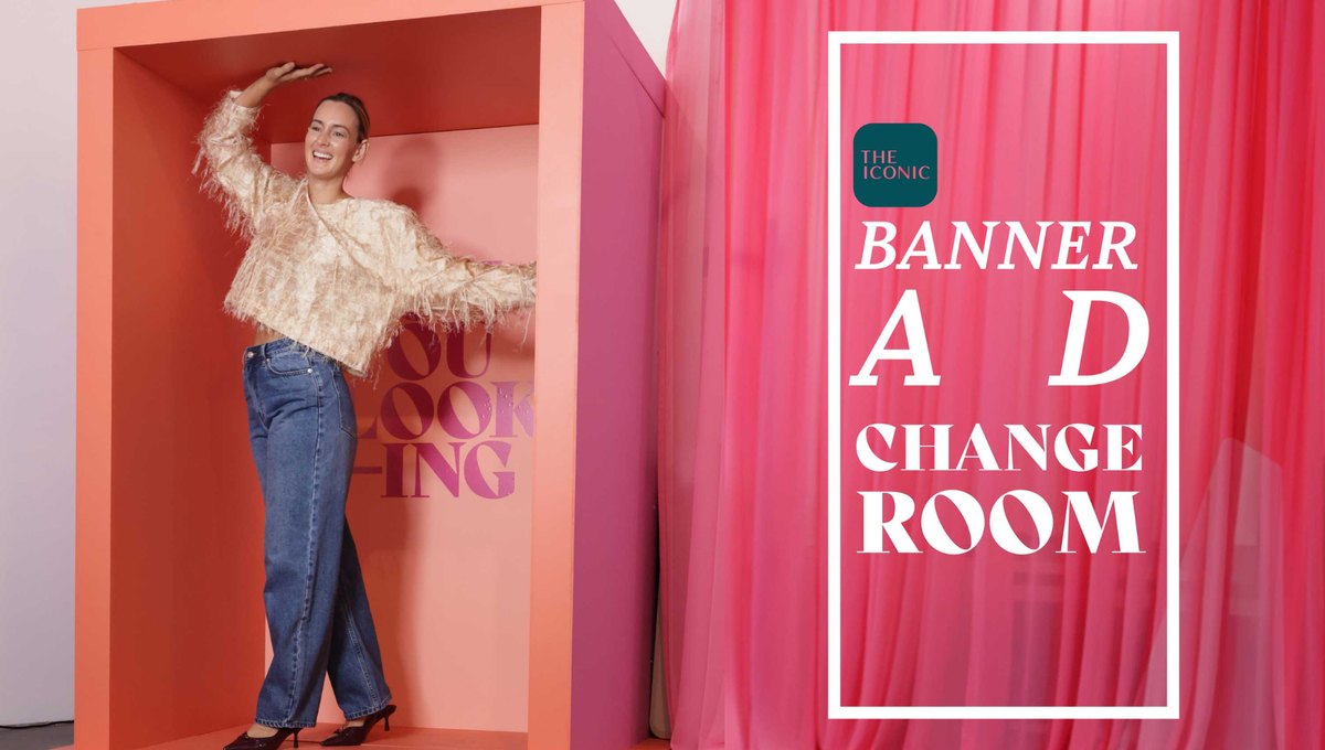 A woman in jeans and a fringed top poses inside a coral box shaped like a banner ad, next to a pink curtain with the words Banner Ad Change Room and THE ICONIC logo
