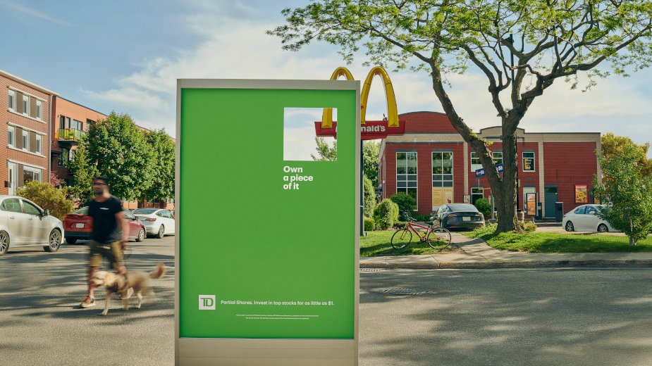 A green TD street panel with a square window cut out that frames part of the McDonald's golden arches behind it, with the line Own a piece of it