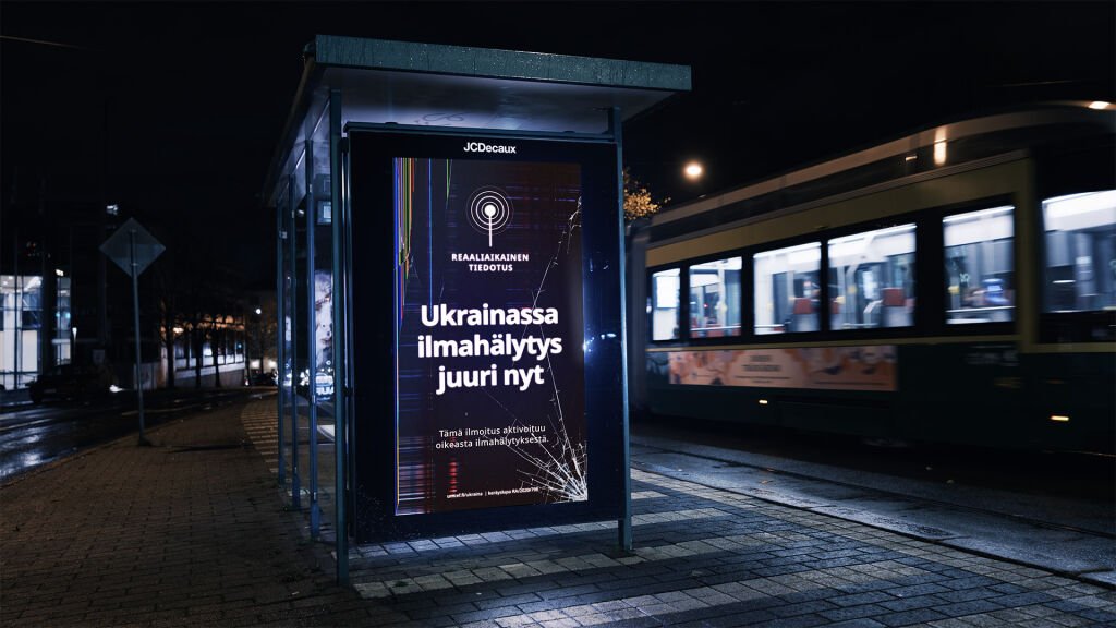 A night-time bus shelter screen in Helsinki reading, in Finnish, air raid alert in Ukraine right now, with a cracked-glass effect, as a tram passes
