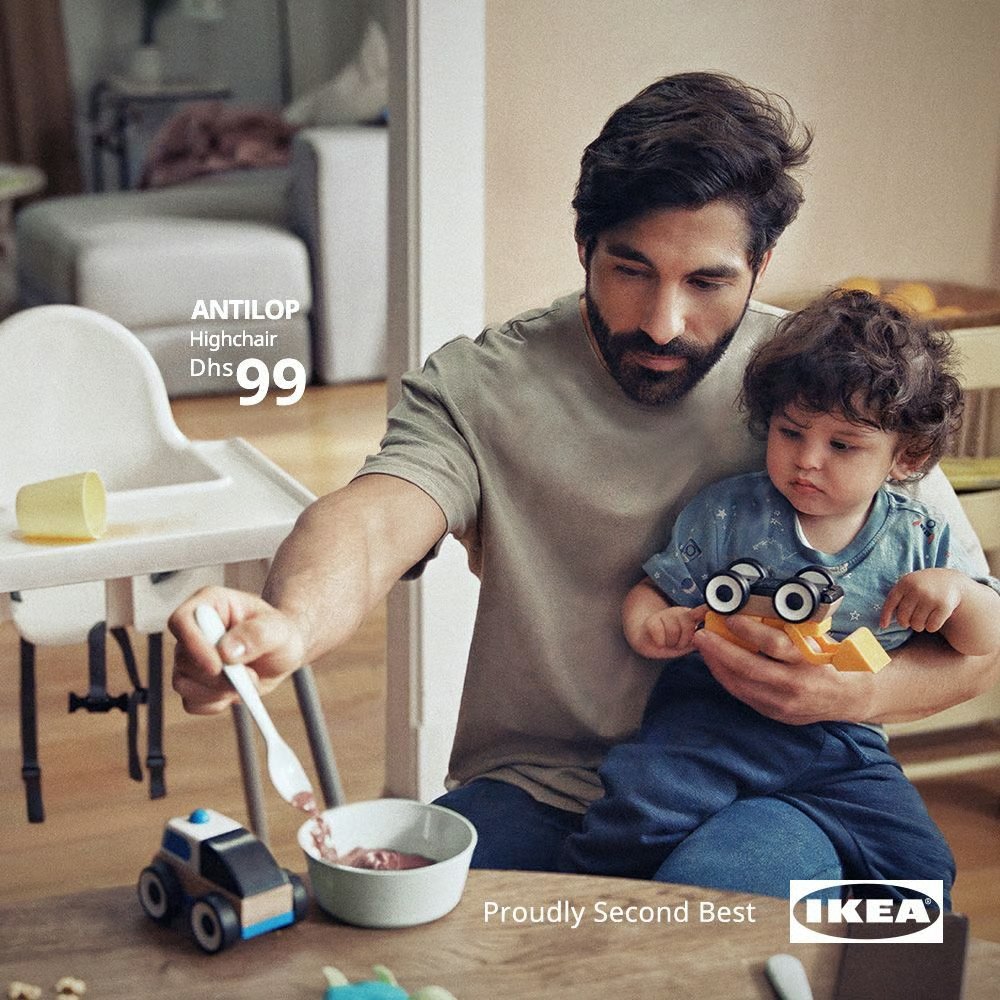 An IKEA Proudly Second Best print ad showing a father feeding his child on his lap while the ANTILOP high chair sits empty behind them