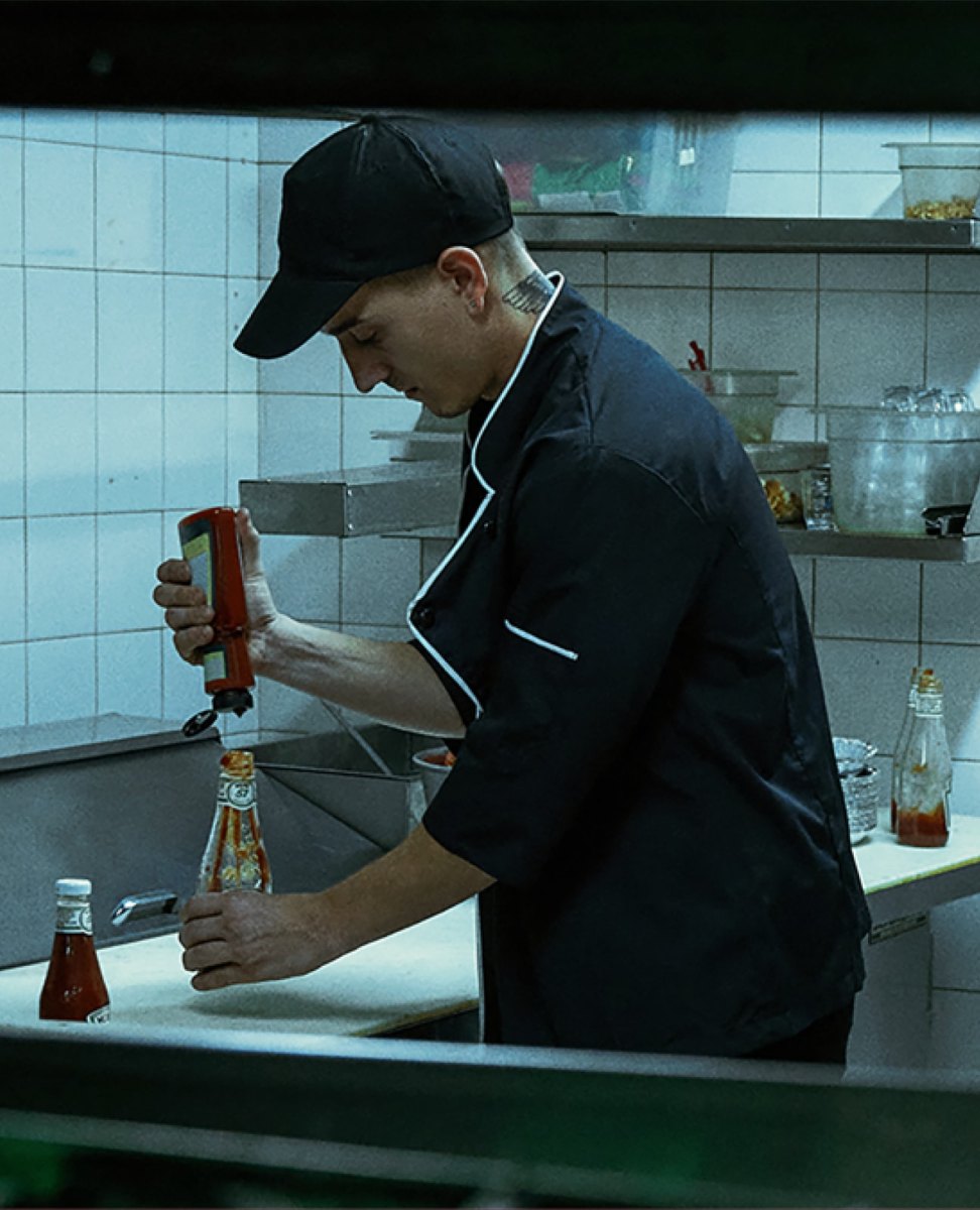 A Heinz Ketchup Fraud campaign photo showing a kitchen worker refilling a glass Heinz bottle from a generic squeeze bottle