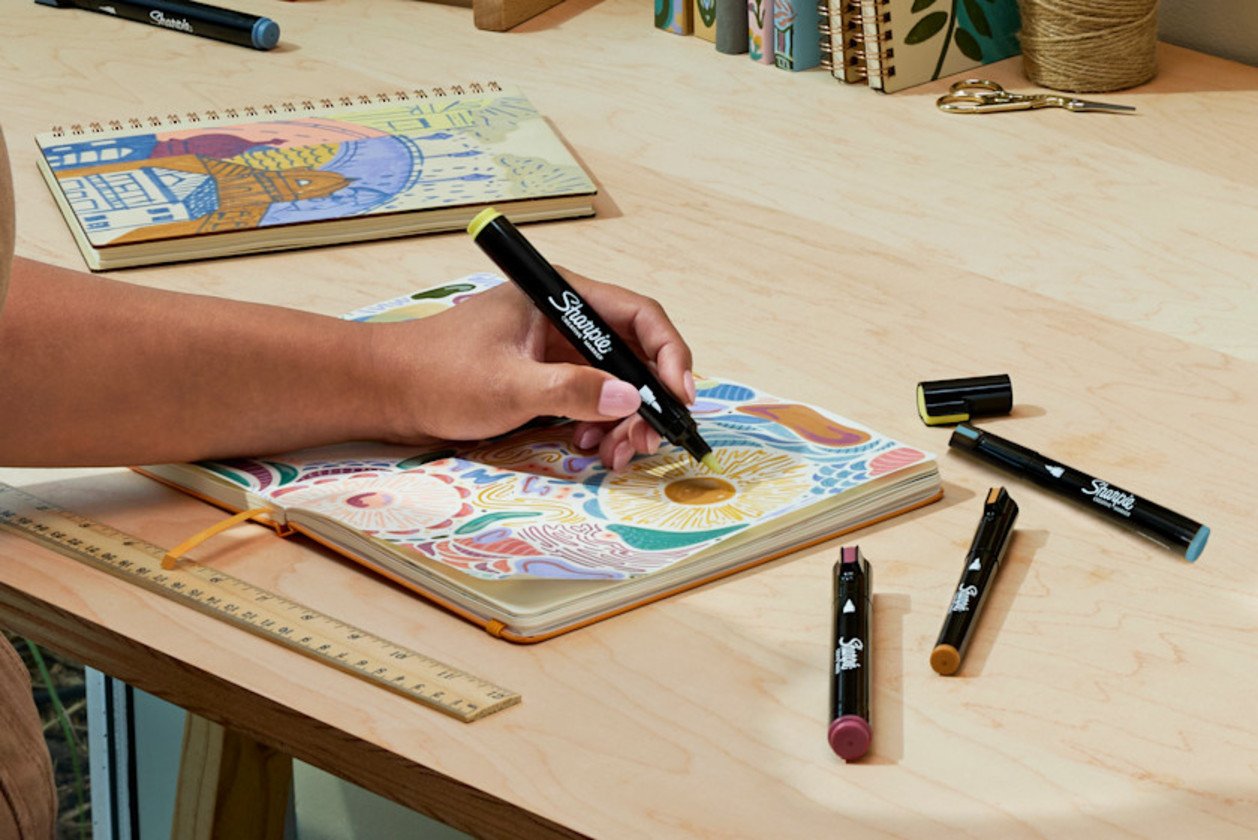 A hand coloring a detailed illustration in a notebook with a Sharpie Creative Marker, other markers on the desk