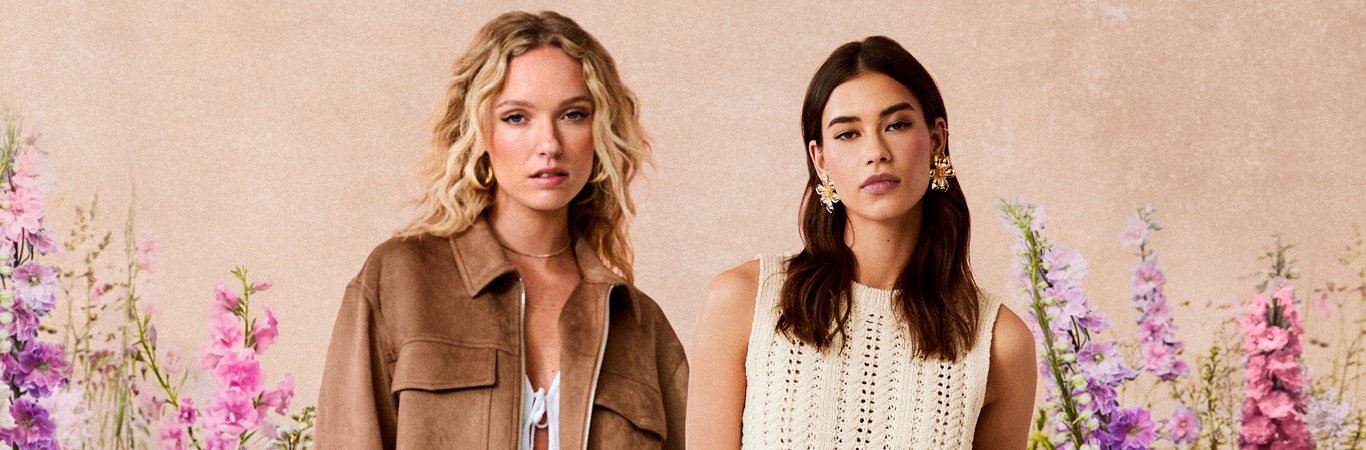 Two models in a suede jacket and a crochet top in front of a beige wall framed by pink and purple flowers