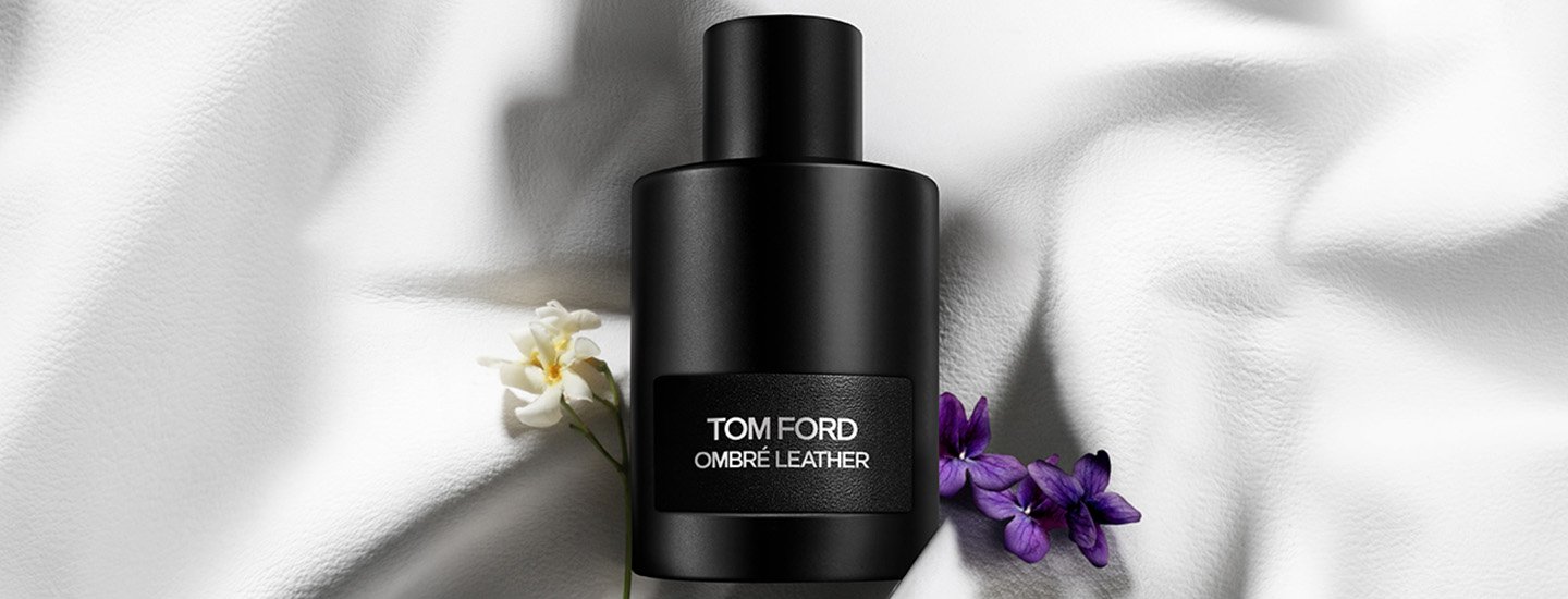 A black Tom Ford Ombre Leather fragrance bottle on a crumpled white surface with a white flower and violets beside it