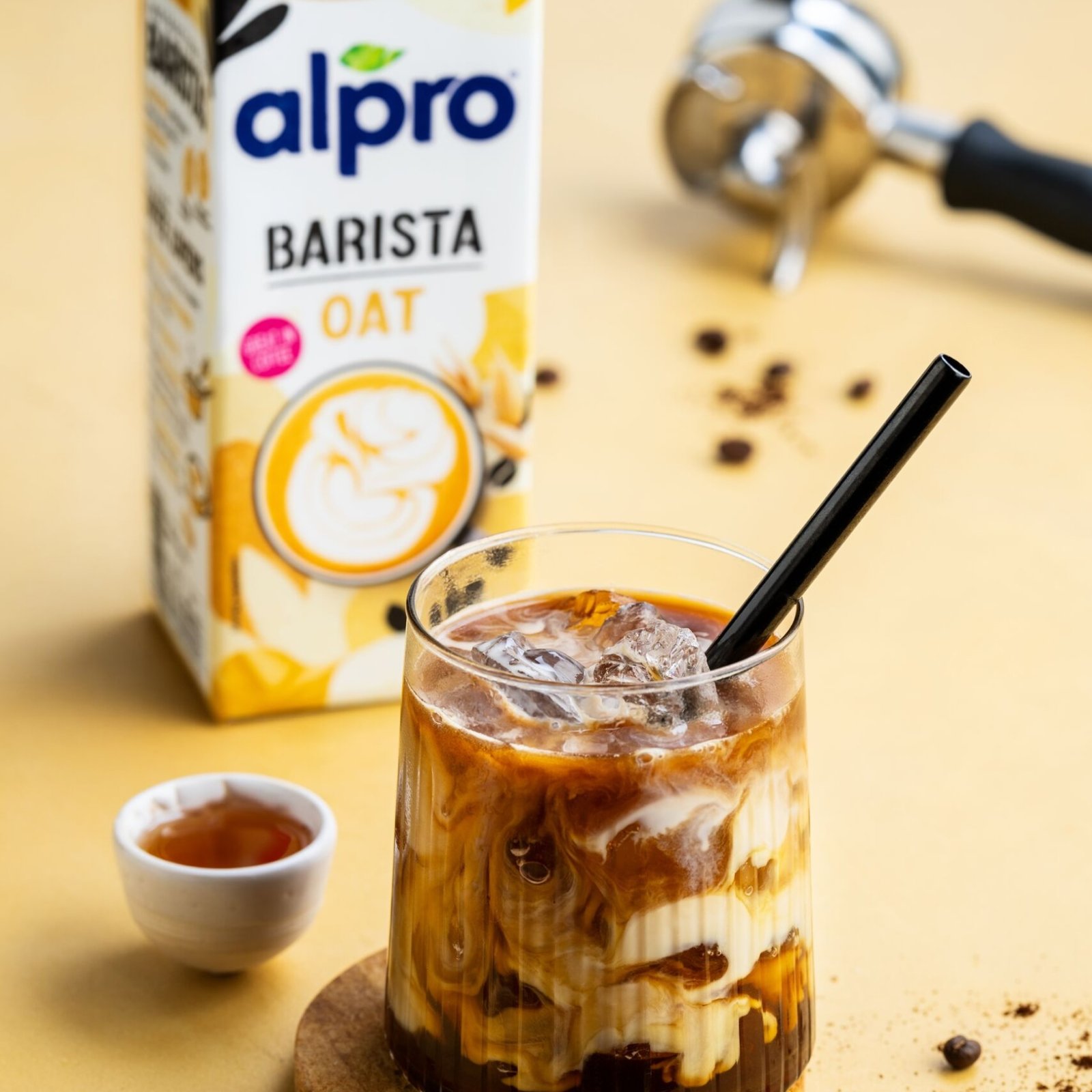 A carton of Alpro Barista Oat next to an iced coffee with oat milk swirling into it