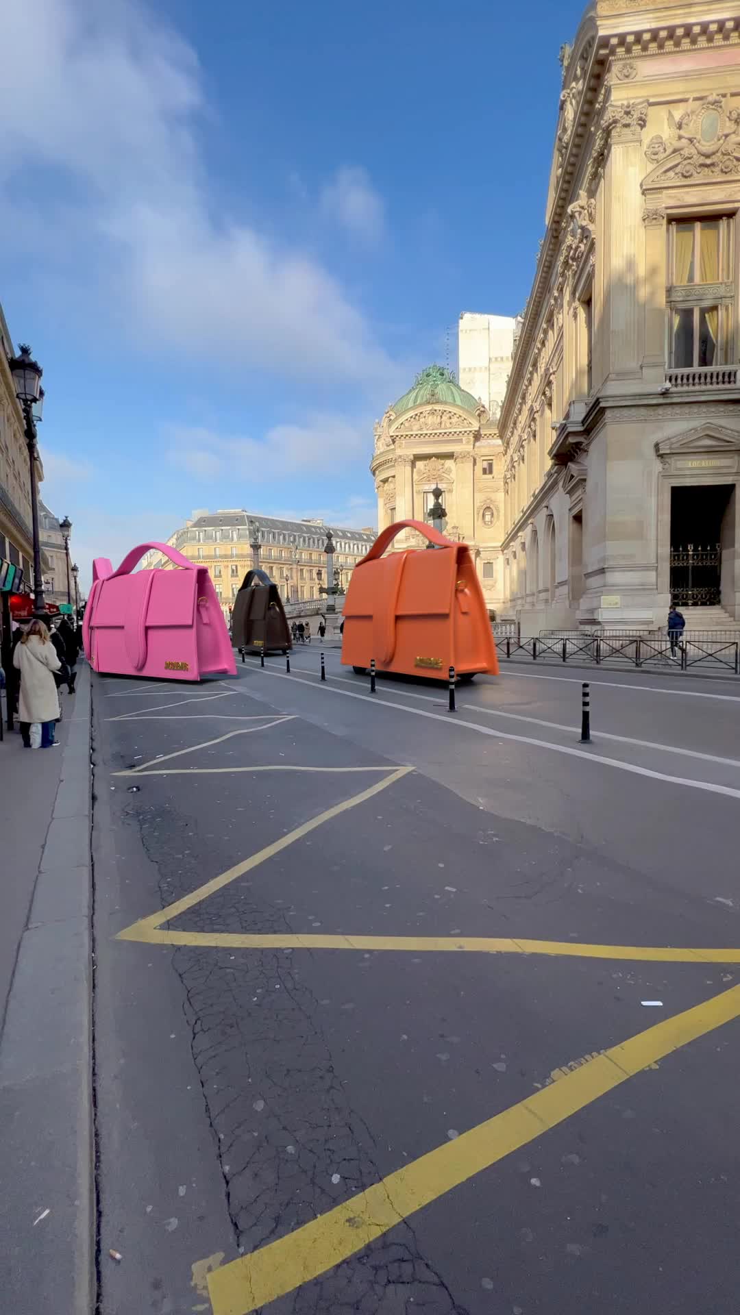 Giant pink, brown and orange Jacquemus Bambino bags driving down a Paris street past the Opera Garnier