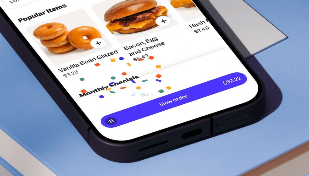 Close-up of a phone showing a Grubhub menu with donuts and a bacon egg and cheese sandwich, with colored confetti bursting above the purple View order button